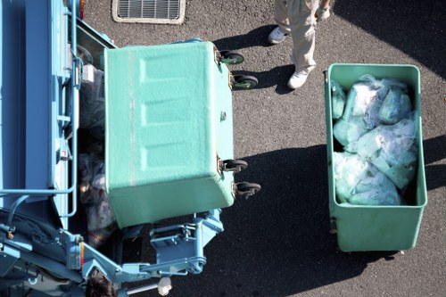 An overhead view of a green waste collection bin filled with clear plastic bags containing rubbish, positioned on a paved outdoor area. Adjacent to the bin is a larger blue waste container with a closed lid, and a person wearing light-colored trousers and white shoes is partially visible standing nearby. The surrounding surface is dark asphalt, indicating a driveway or street, and the scene appears to be part of a rubbish removal operation in a residential or commercial area, possibly within the New Cross postcode region. The image reflects typical waste disposal practices handled by Office Clearance New Cross, highlighting their rubbish collection service.