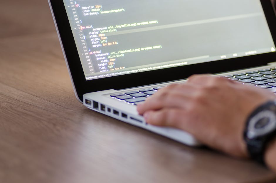 Close-up image of a person's right hand operating a silver laptop with a black keyboard. The hand is positioned over the touchpad, and the individual is wearing a wristwatch with a black strap and a r