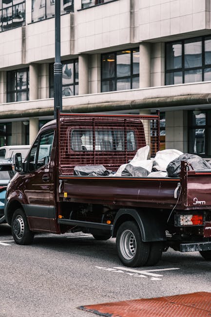 A small burgundy flatbed truck parked on a city street in front of a modern office building with large glass windows. The truck's cargo area is filled with various rubbish bags and scattered debris, i