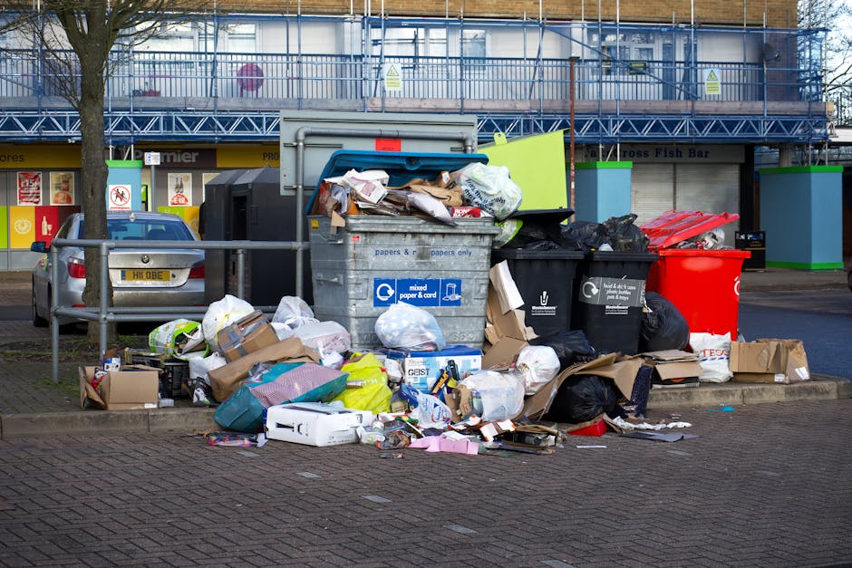A collection of discarded household items and debris piled on a paved outdoor surface, including wooden garden furniture with slatted backs and legs leaning against a wall, a partially damaged broken 